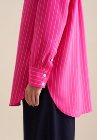 Fuchsia gestreepte blouse met een afgeronde zoom, knoopmanchetten en een losse pasvorm; gemaakt van een lichte stof met witte verticale strepen.