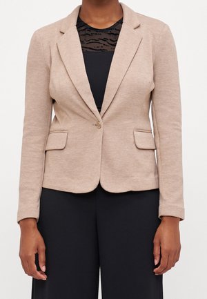 Femme portant un blazer beige à un bouton sur un haut noir avec empiècement transparent à motifs et un pantalon noir, debout devant un fond uni.