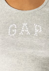 La camiseta sin mangas de canalé gris presenta el logo de "GAP" adornado con diseños de flores en blanco. La tela tiene una apariencia suave y texturizada.