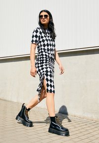 Abito in bianco e nero con motivo a diamante, dettagli arricciati, spacco laterale, abbinato a stivali neri con tacco alto e lacci. Ambientazione esterna illuminata dal sole.