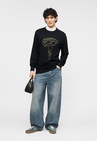 Μαύρο φούτερ με χρυστό σχέδιο λουλουδιού, συνδυασμένο με φαρδιά μπλε τζιν και πράσινα αθλητικά παπούτσια. Το μοντέλο κρατάει μια μικρή μαύρη τσάντα χειρός.