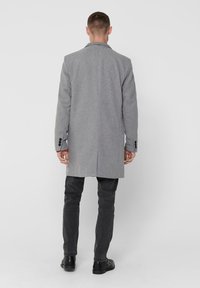 Grau melierter Wollmischmantel mit langer Schnittführung, einer Rückenschlitz und schwarzen Knöpfen an den Manschetten. Kombiniert mit grauen Hosen und schwarzen Schuhen.