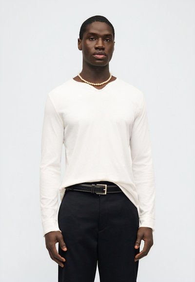 Άνδρας που φοράει λευκό πουκάμισο με μακριά μανίκια, μαύρο παντελόνι με ζώνη και μονόκλωνο περιδέραιο με μαργαριτάρια, στέκεται μπροστά σε απλό ανοιχτό φόντο.