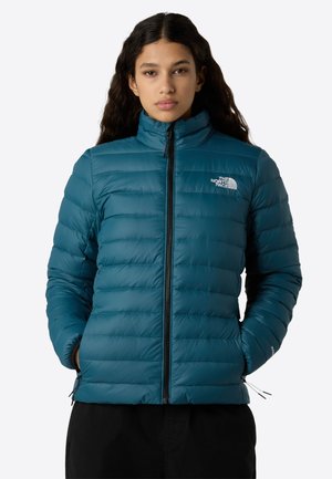 Femme portant une veste matelassée teal de North Face, les mains dans les poches, se tenant devant un fond blanc uni.