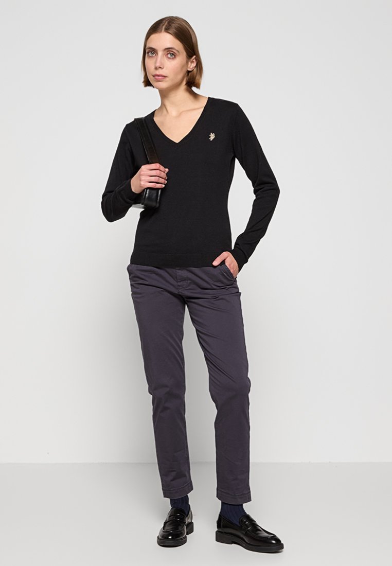 Jeune femme portant un pull noir en V, un pantalon gris foncé, des mocassins noirs, tenant un sac à main noir, se tenant debout contre un mur clair.