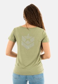 T-shirt à manches courtes de couleur vert olive avec un décolleté décontracté, présentant un design en étoile argentée et en chevrons au dos. Matériau en mélange de coton.