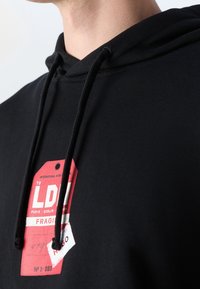 Gros plan d'une personne portant un sweat à capuche noir avec des cordons noirs et une étiquette graphique rouge indiquant "LDN FRAGILE" sur la poitrine.