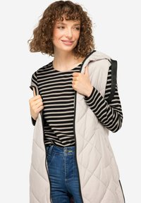 Lichte beige gewatteerde vest met een ritssluiting aan de voorkant, gedragen over een zwart-beige gestreepte shirt; voorzien van een capuchon en zijzakken.