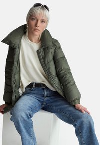 Oliwkowa puchowa kurtka z dużym kołnierzem; kremowy sweter pod spodem; wąskie niebieskie dżinsy. Białe tło, jednolity kolor, teksturowana tkanina.
