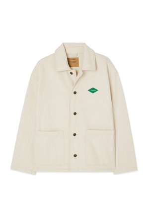Veste couleur crème en tissu robuste, avec un col pointu, une fermeture à boutons, deux poches avant et un logo en forme de diamant vert sur la poitrine gauche.