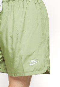 Světlé zelené šortky Nike s elastickým pasem, šňůrkou na stažení, bočními kapsami a bílým logem na dolním levém rohu. Hladká textilie.