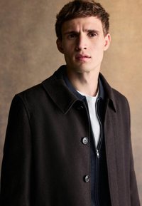 Brązowy płaszcz z mieszanki wełny z zaostrzonym kołnierzem, z dwoma guzikami, nałożony na granatową kurtkę zapinaną na zamek i białą koszulkę z okrągłym dekoltem.