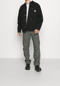 Czarna kurtka typu canvas z zatrzaskami i dwoma kieszeniami na piersi, zestawiona z luźnymi oliwkowymi spodniami cargo i czarnymi skórzanymi botkami.
