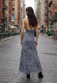 Šedé květované maxi šaty s tenkými ramínky, zavazováním vzadu a splývavou siluetou. Viditelná černá kotníková obuv. Městské pozadí.
