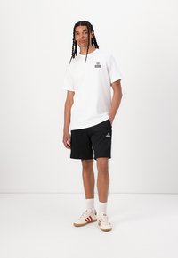 White short-sleeve t-shirt featuring a subtle logo on the chest, paired with black shorts and red-striped sneakers. Smooth fabric texture.