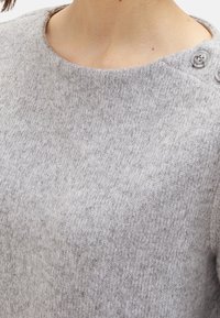 Šedý pletený svetr s kulatým výstřihem, texturovanou tkaninou a knoflíkovými detaily na jednom rameni. Měkký, hřejivý materiál s hladkým povrchem.
