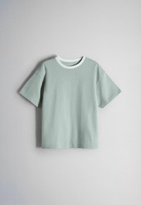 Camiseta de manga corta de color verde claro con cuello redondo blanco colgada contra una pared lisa de color claro.