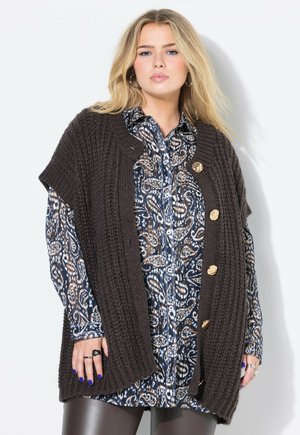 Blonde Frau, die ein gemustertes Hemd mit Knöpfen unter einem grob gestrickten, dunkelbraunen Weste mit goldenen Knöpfen und dunklen Lederhosen trägt.