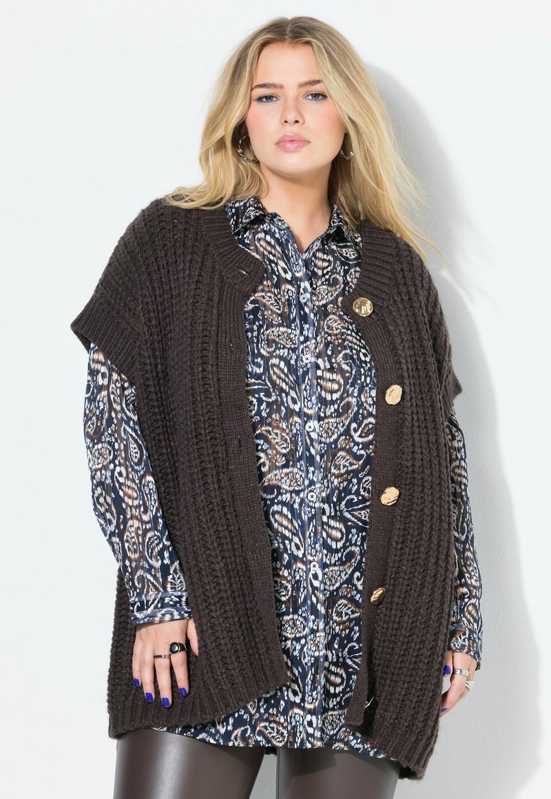Donna bionda che indossa una camicia a maniche lunghe stampata sotto un maglione spesso marrone scuro con bottoni dorati e pantaloni di pelle nera.