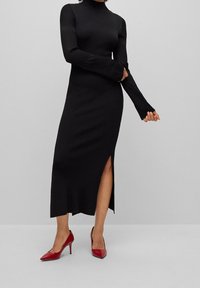 Kvinna som bär en lång svart ribbad klänning med nätärm och sidoflik, i kombination med röda spetsiga klackskor, som står på ett ljust grått golv.