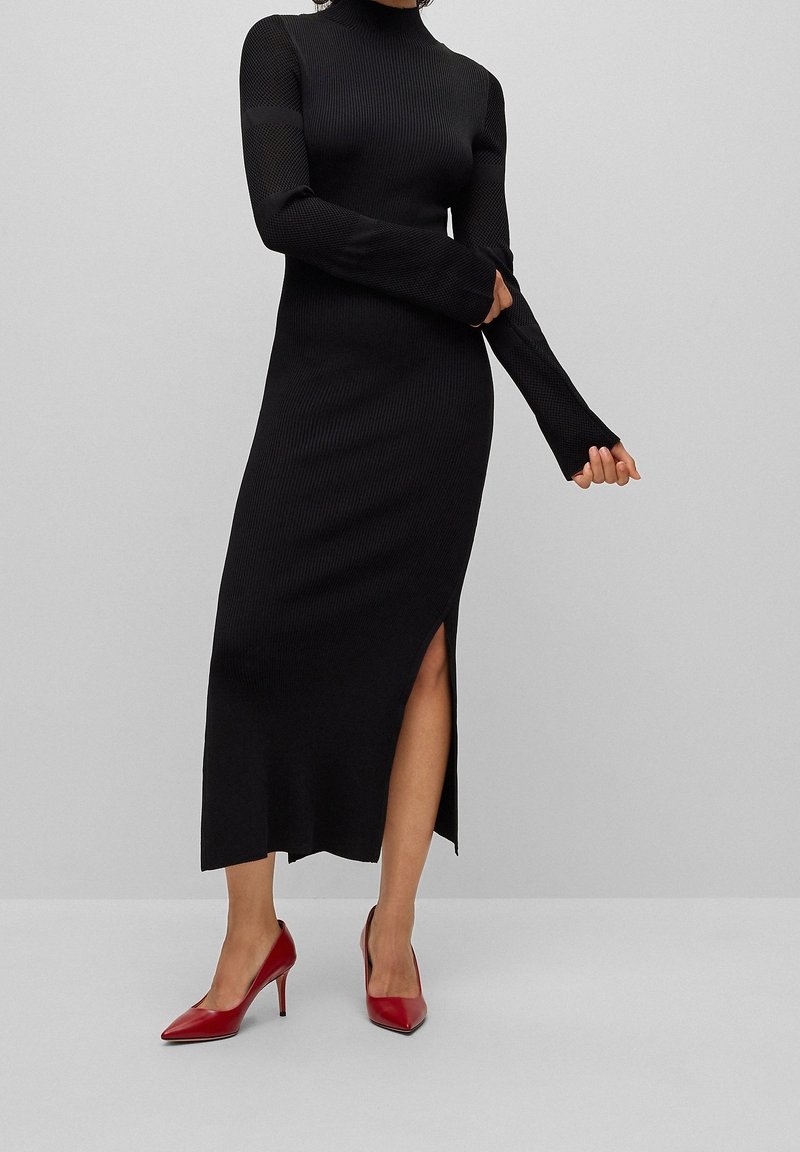 Kvinna som bär en lång svart ribbad klänning med nätärm och sidoflik, i kombination med röda spetsiga klackskor, som står på ett ljust grått golv.