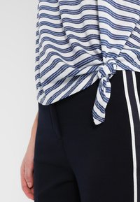 Person med en blå och vit randig skjorta knuten i midjan och mörka byxor med vita sidor, hand vilande vid höften.