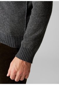 Šedý vlněný svetr s žebrovanými manžetami, který má strukturovaný povrch. Detailní záběr na ruku odpočívající na rukávu, který zvýrazňuje detaily manžety.