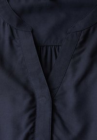 Blusa blu navy con scollatura a V, tessuto morbido e dettagli arricciati sotto il collo con bordi cuciti sottili.