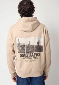 Sudadera beige con un estampado gráfico de cáctus en el Parque Nacional Saguaro, con letras grandes y un corte holgado. Textura suave, diseño tipo pullover.
