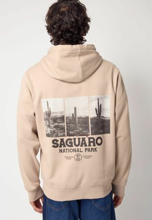 Sudadera beige con un estampado gráfico de cáctus en el Parque Nacional Saguaro, con letras grandes y un corte holgado. Textura suave, diseño tipo pullover.