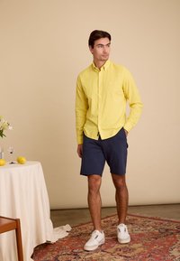 Geel overhemd met lange mouwen, marineblauwe shorts en witte sneakers. De achtergrond is een neutrale kleur, met een tafel en decoratieve items.