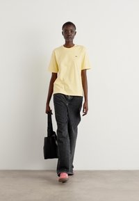 Gul kortärmad T-shirt, svarta vida jeans och rosa skor. Modellen bär en svart tote-väska. Minimal bakgrund.