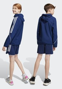 Marineblauwe sportjas en short set met witte strepen en accenten. Lichtgewicht stof met een ontspannen pasvorm en capuchon. Schoenen in gemengde kleuren.