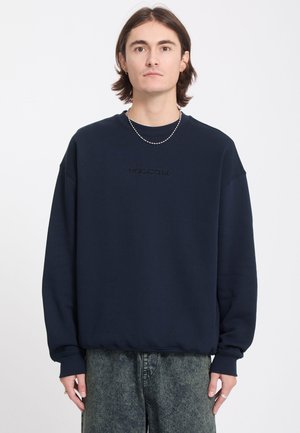 Jeune personne aux cheveux bruns mi-longs, portant un sweat-shirt Volcom bleu marine et un jean gris, debout devant un fond blanc.