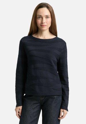 Woman with long brown hair wearing a dark navy textured long-sleeve sweater and dark jeans, standing against a plain background.