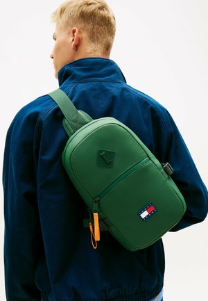 Blond persoon in een donkerblauw jack met een groene schoudertas met voorvak met rits en een Tommy Jeans logo.