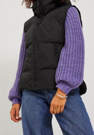 Persona con un chaleco negro acolchado sobre un suéter grueso de lana púrpura con mangas abullonadas y jeans azules, con una pulsera plateada en la muñeca.