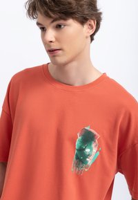 T-shirt di cotone in arancio ruggine con collo a giro. Presenta una stampa grafica verde e rossa sul lato sinistro. Maniche corte, vestibilità rilassata.