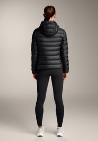 Črna puhovka s kapuco, quilted tekstura in elastični manšeti, v kombinaciji s črnimi leginami in belimi supergami, prikazana s hrbta.