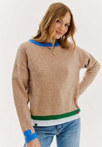 Beżowy sweter z niebieskimi i zielonymi prążkowymi akcentami przy kołnierzu i krawędzi, o luźnym kroju i miękkiej teksturze. Długie rękawy z kolorowym wykończeniem.