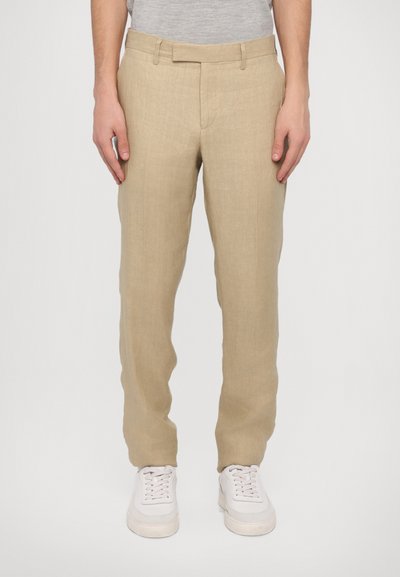 Pantaloni in lino beige con taglio dritto, dotati di vita piana, passanti per cintura e una texture liscia. Abbinati a sneakers bianche.