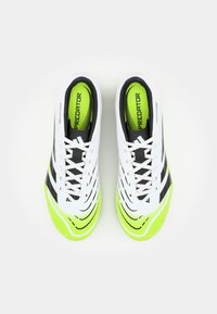 Chaussures de football blanches avec des accents noirs, des semelles vertes et un design texturé. Présentent un système de laçage et une étiquette « PREDATOR » bien visible à l'intérieur.
