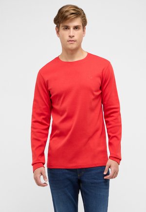 Young man with light brown hair wearing a plain red long-sleeve shirt and blue jeans, standing against a white background.