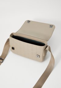 Borsa rettangolare beige con un motivo geometrico testurizzato. Presenta una chiusura magnetica, hardware argentato e una tracolla regolabile. L'interno è scuro.