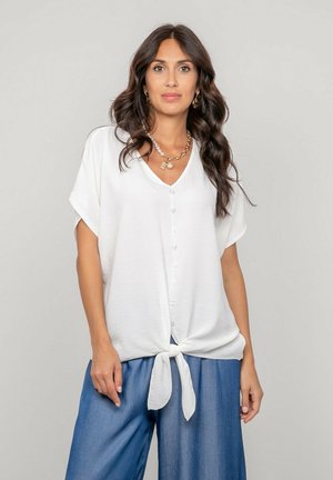 Femme aux longs cheveux bruns portant un chemisier blanc boutonné noué à la taille et un pantalon bleu ample, debout devant un fond uni.