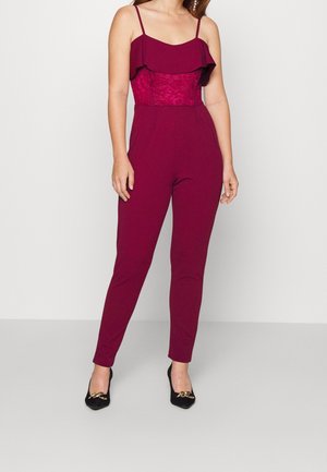 Bordeaux jumpsuit met een kanten bovenstuk, gerimpelde halslijn, getailleerd silhouet en smal toelopende broekspijpen, gecombineerd met zwart versierde flats.