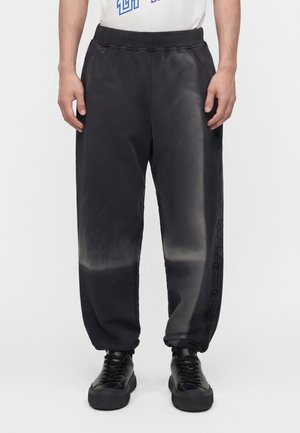 Homme portant un pantalon de jogging tie-dye noir avec des poignets élastiques et des baskets noires, debout devant un fond blanc.