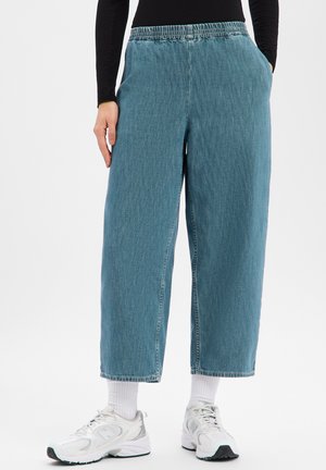 Hellblaue Denim-Hose mit weitem Bein, elastischem Bund, seitlichen Taschen und roher Saumkante. Kombiniert mit weißen Sportschuhen und Socken.