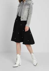 Szara dżinsowa kurtka noszona na szarym topie, zestawiona z czarną rozkloszowaną spódnicą ozdobioną białymi perłami oraz błyszczącymi białymi botkami za kostkę z sznurowadłami.