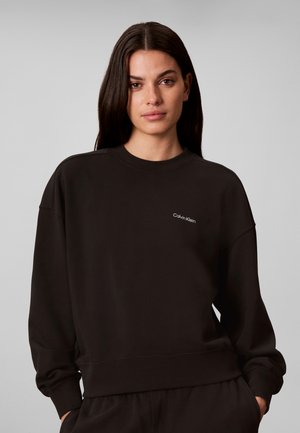 Kobieta z długimi ciemnymi włosami, ubrana w czarną bluzę Calvin Klein oraz dopasowane spodnie na jednolitym szarym tle.